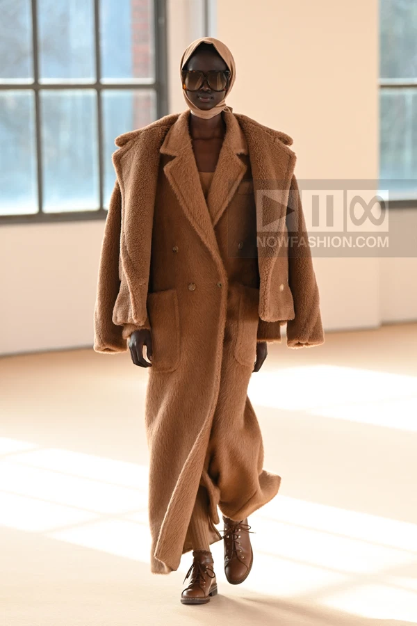 A person wearing a long camel coat, a matching jacket draped over shoulders, a headscarf, and brown lace-up boots.
