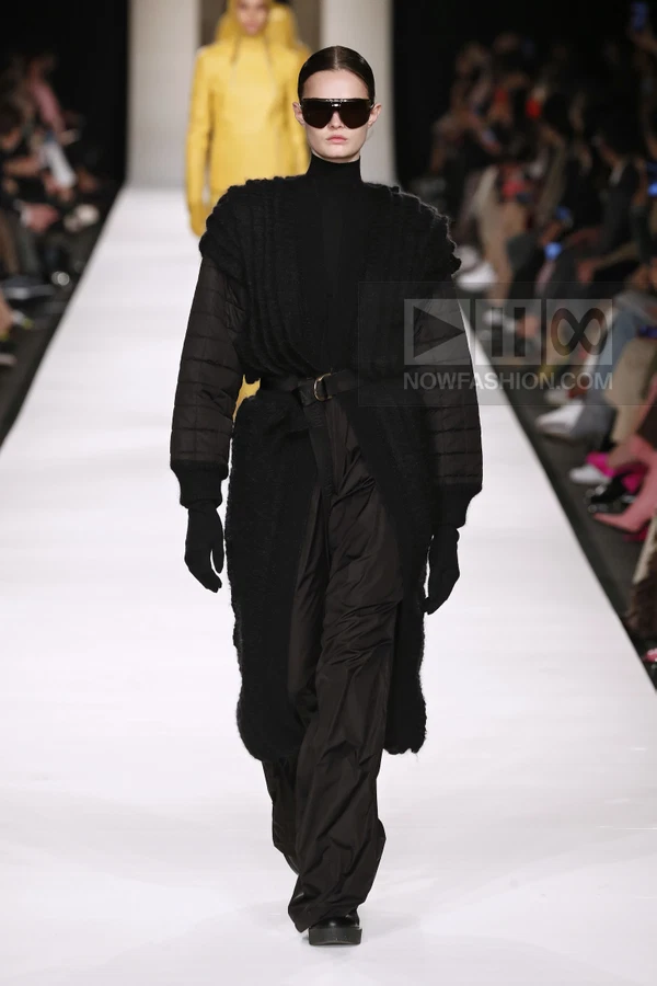 A person walks down a runway wearing a black quilted coat, black turtleneck, and black trousers, with dark sunglasses.