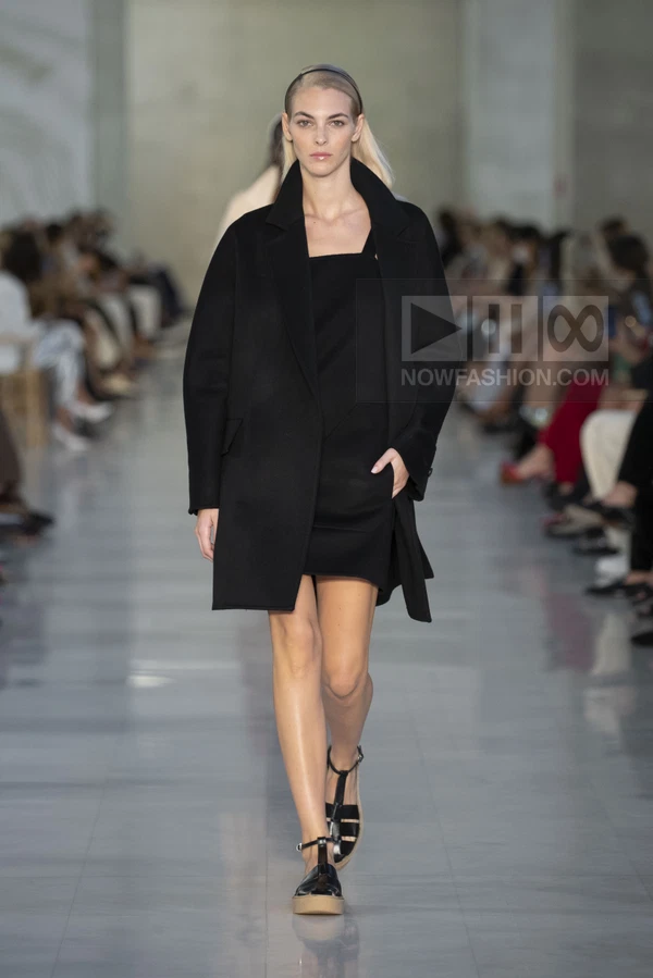 A model walks the runway in a black coat over a matching mini dress and flat sandals.