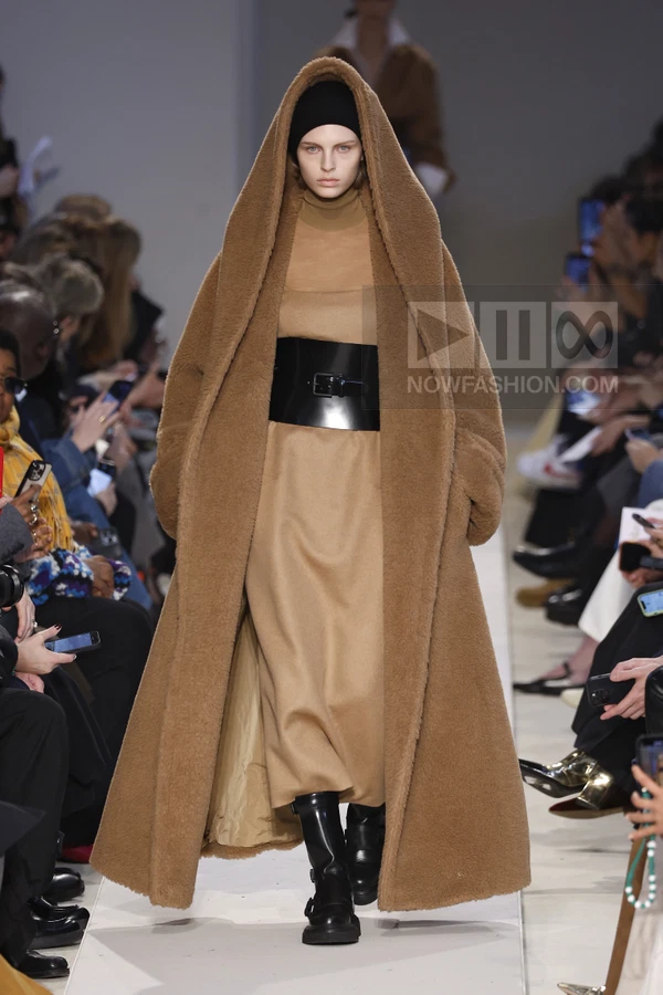 Model wearing a long brown hooded faux-fur coat over a camel turtleneck dress with a wide black patent leather waist belt.