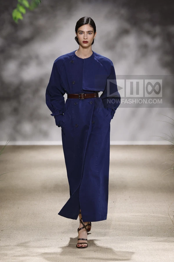 Model wearing a navy blue long coat with buckled cuffs and a brown leather waist belt on a runway.