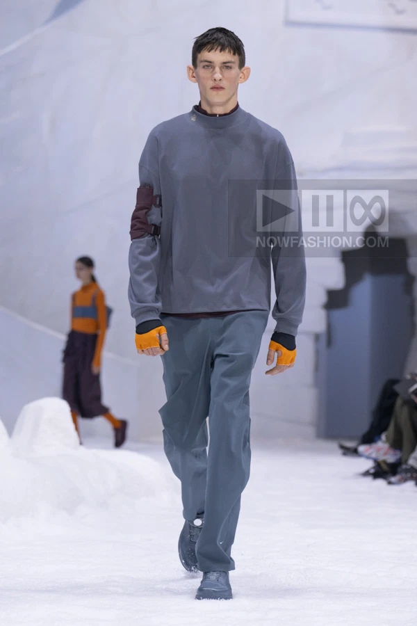 A person walking on a white runway wearing a gray long-sleeve top and matching trousers with orange fingerless gloves.