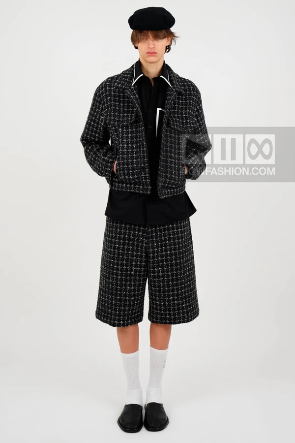 A person wearing a black and white tweed jacket and matching shorts, a black shirt, a beret, white socks, and black shoes.