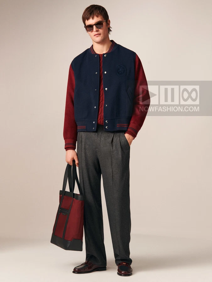 A man wearing a navy and burgundy varsity jacket, cable-knit sweater, and gray trousers, holding a burgundy tote bag.