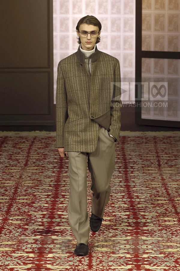 Model wearing a brown plaid jacket, gray turtleneck, and wide-leg trousers on a red patterned runway carpet.