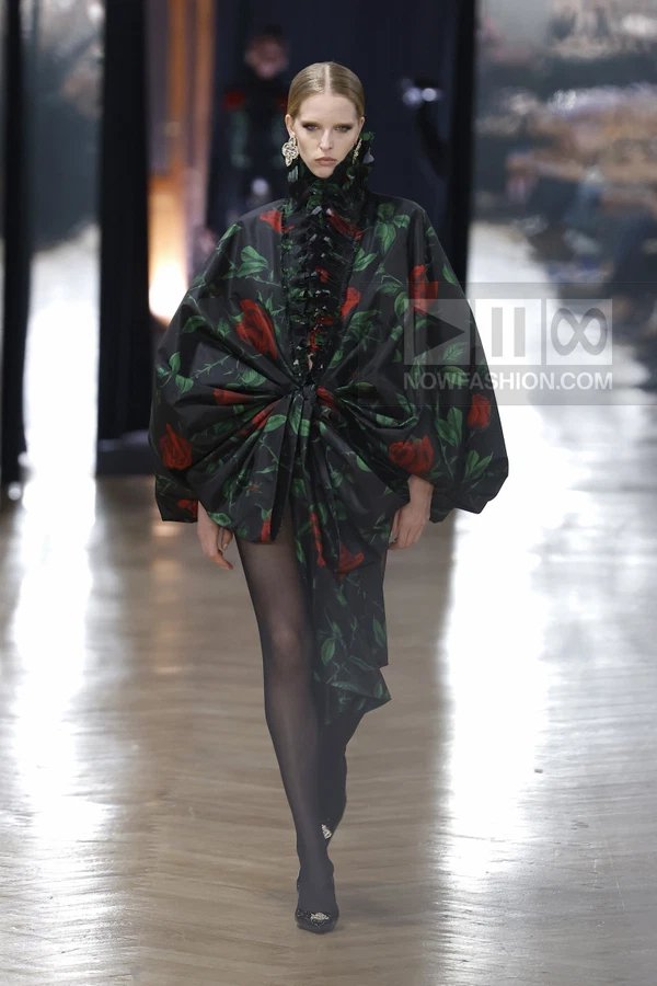 A model walks the runway wearing a black oversized floral print jacket with a ruffled collar and sheer black tights.
