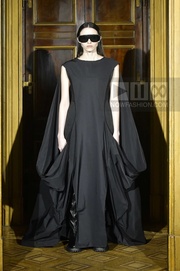 A person wearing a black floor-length sleeveless dress with draped side panels and black boots, standing in a doorway.