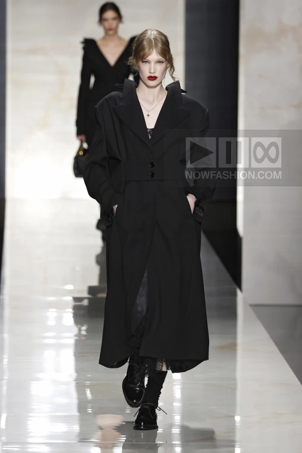 Model walking in a black wool coat with wide notched lapels, cinched waist, and lace hem, wearing black lace-up shoes.