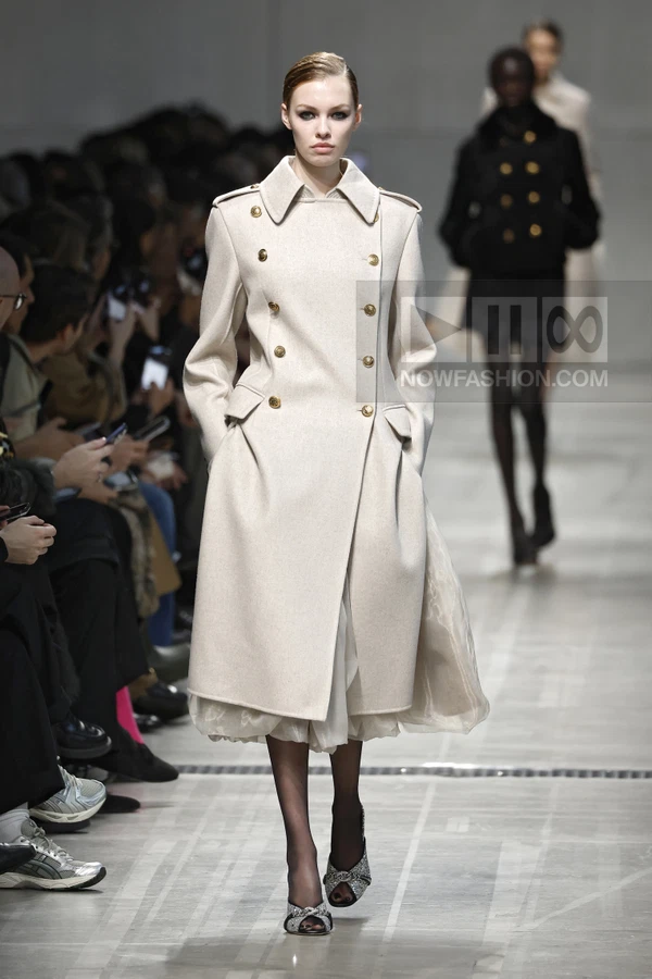 Model walking in a beige double-breasted wool coat over a sheer skirt, with black tights and embellished open-toe heels.