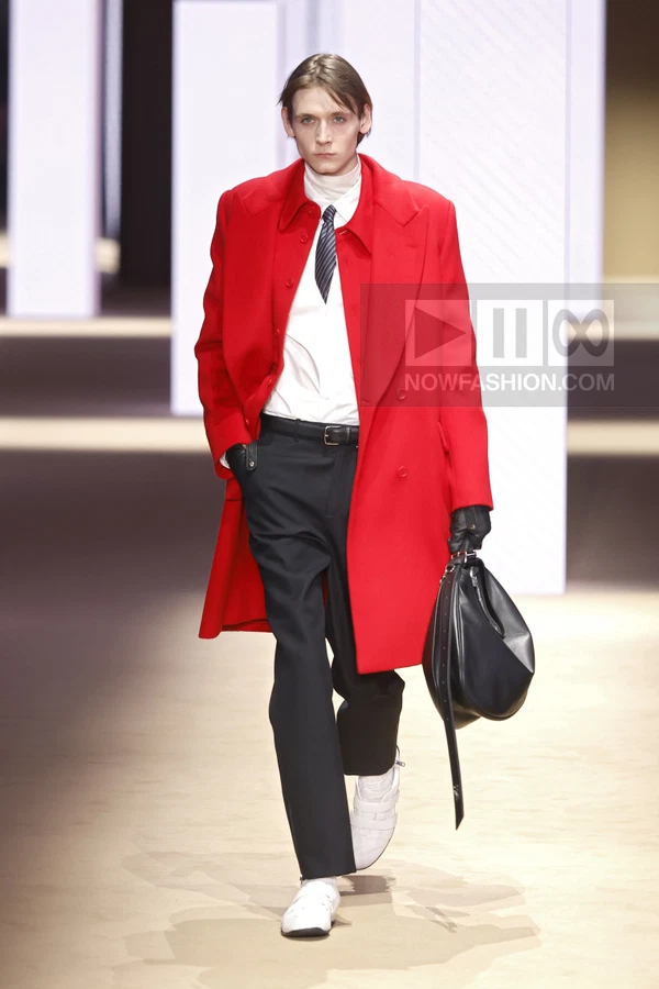 Model walks in a bright red wool coat over a white shirt, striped tie, and black trousers with white sneakers.