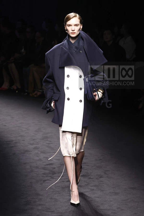 Model walking in a navy and white double-breasted coat, light skirt with ties, and navy shoulder bag on a dark runway.