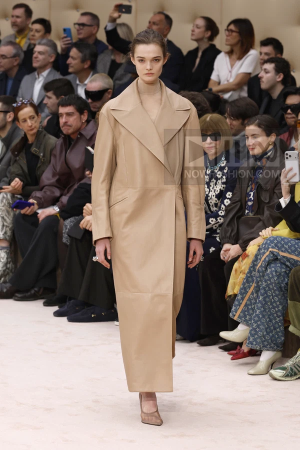 A person walks a runway wearing a long, tan leather coat with a high collar and matching pointed-toe heels.
