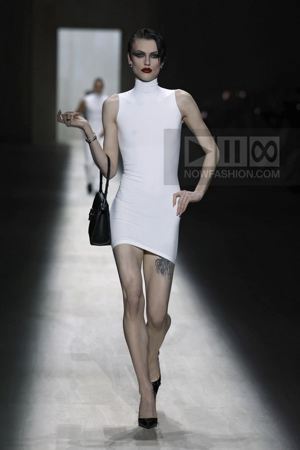 Model walking in a white sleeveless mini dress with a high neck, carrying a black top-handle bag on a runway.