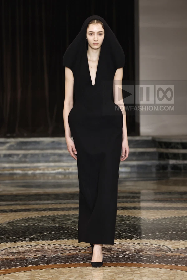 Model wearing a black hooded floor-length dress with a deep v-neckline and structured hip silhouette on a runway.