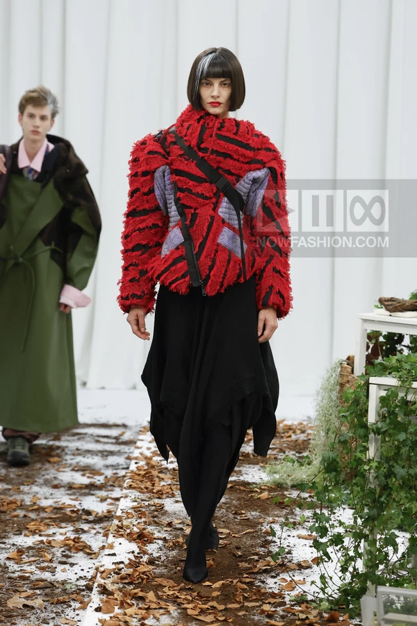 Model wearing a red and black textured jacket with a black skirt on a runway covered in autumn leaves.