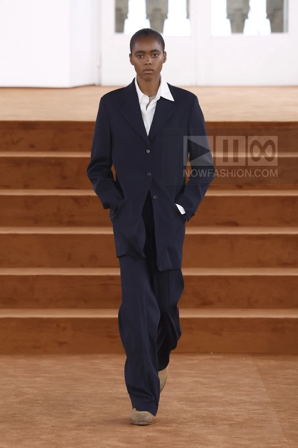 A person walks on a runway wearing a dark navy suit with a white shirt and tan shoes.