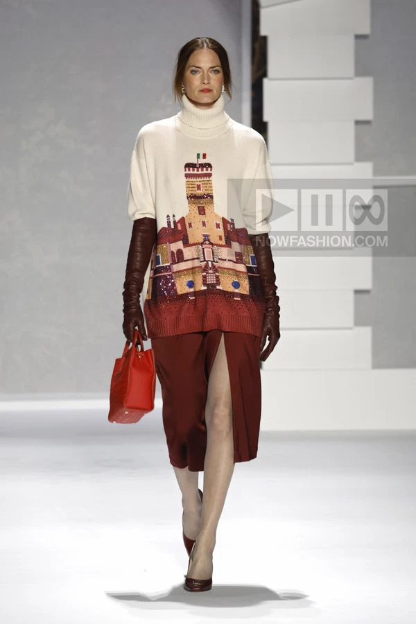 Model walking in a cream knit sweater with a castle graphic, dark red midi skirt with a front slit, and leather gloves.