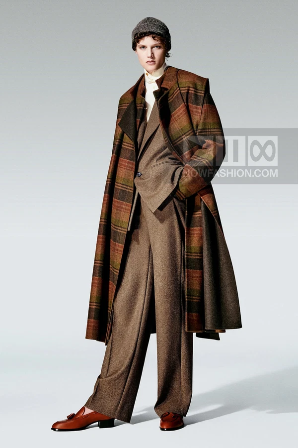 Model wearing a plaid wool coat over a brown suit, cream shirt, and beanie, standing against a plain gray background.