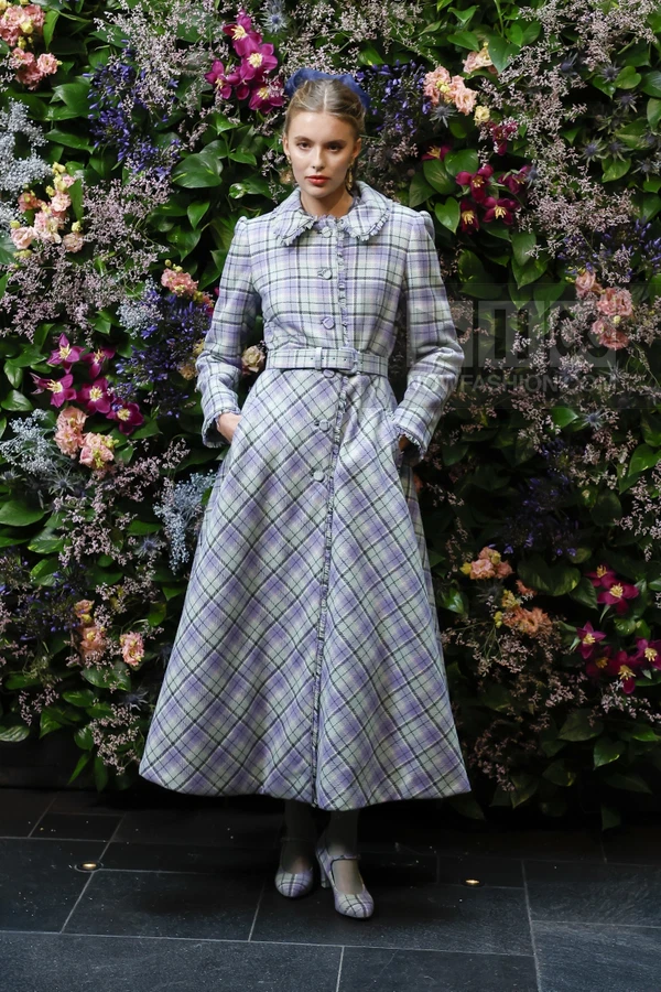 A person wearing a long, plaid, belted coat with a ruffled collar, standing in front of a wall of flowers.