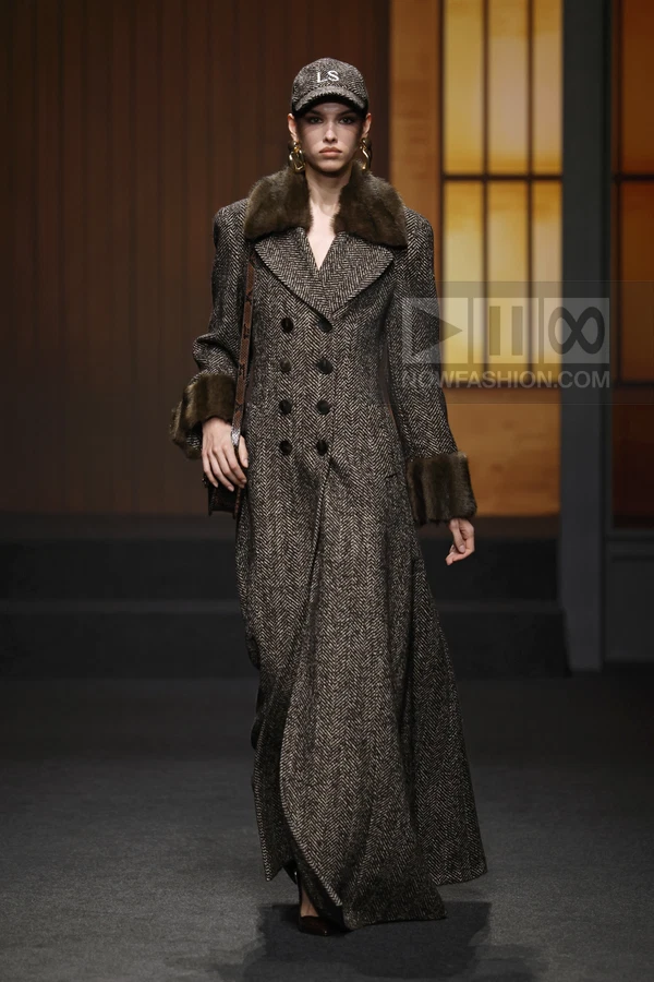 Model wearing a long brown herringbone double-breasted coat with fur collar and cuffs, paired with a baseball cap.