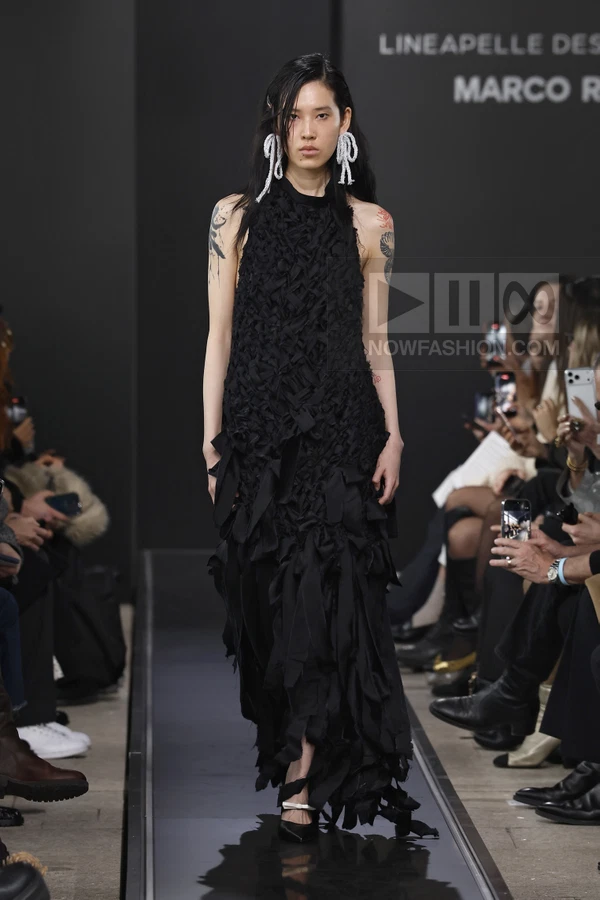 A model walks the runway in a black sleeveless textured maxi dress with fringed hem details and white bow earrings.