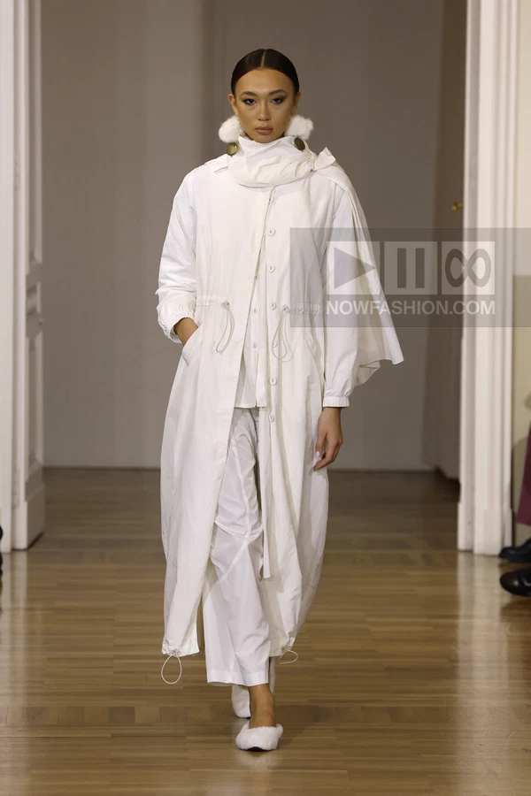 A person wearing a white long coat with a drawstring waist and white trousers, walking on a wooden floor.