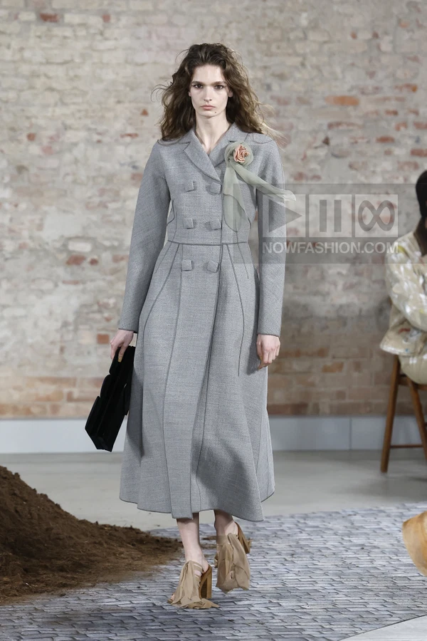 A model walks in a gray wool coat with a rose pin and sheer scarf, holding a black clutch and wearing tan heeled sandals.