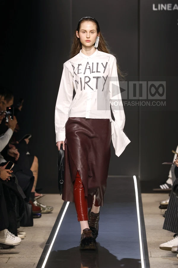 Model on runway wearing a white shirt with text, a dark red leather skirt over red patent trousers, and dark shoes.