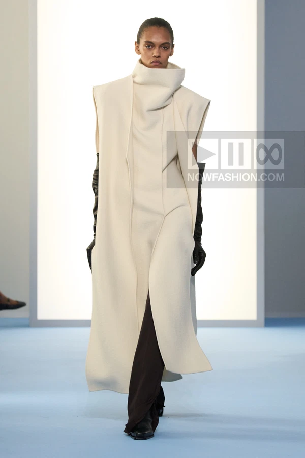 A model walks the runway in a long cream sleeveless coat over dark brown trousers and black leather opera gloves.