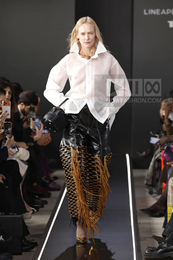 Model on runway wearing a white sheer button-down shirt and black leather pants with tan fringe and woven details.
