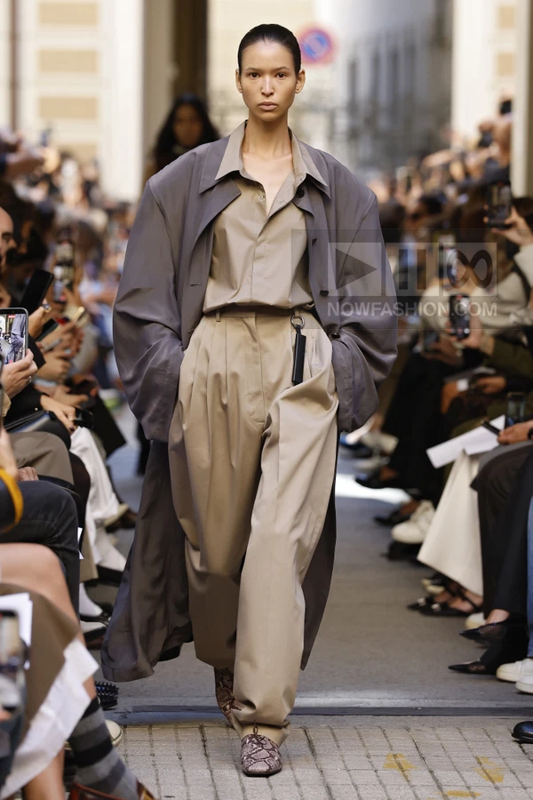 Model walking in a taupe oversized coat, beige button-down shirt, and pleated trousers with snakeskin-print shoes.