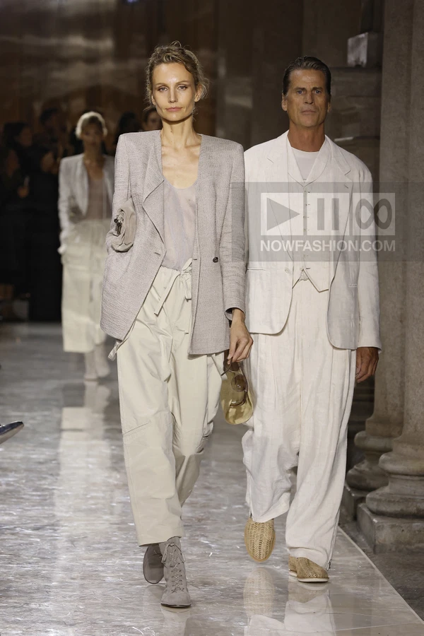 Two models walking on a runway wearing neutral-toned tailored jackets, trousers, and light-colored footwear.