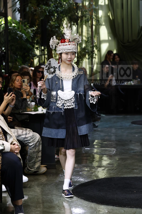 A person wearing a denim jacket, pleated skirt, and elaborate silver headwear, holding a hand fan on a runway.