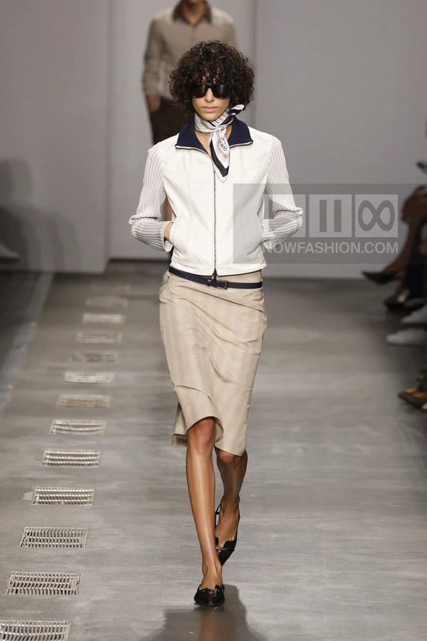 A model walks the runway wearing a white zip-up jacket, a beige plaid midi skirt, and dark sunglasses.