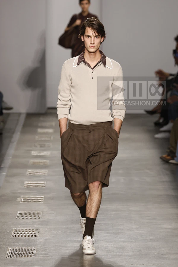 A person walks down a runway wearing a beige long-sleeve polo shirt over a striped shirt and brown pleated shorts.