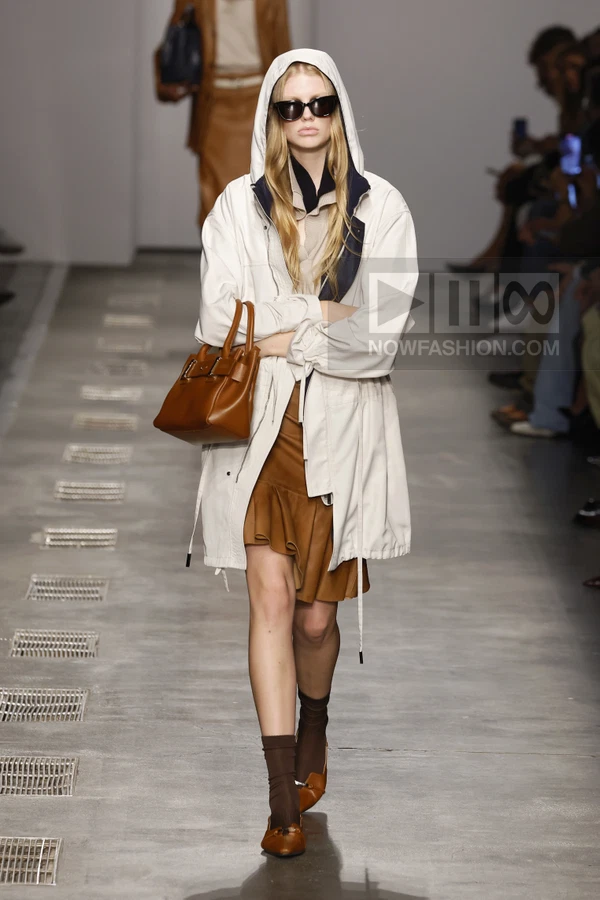 Model wearing a white hooded windbreaker over a brown skirt, carrying a brown top-handle bag on a runway.
