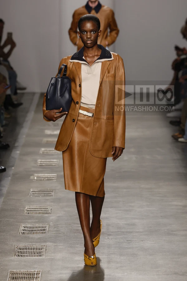 Model walking on runway wearing a tan leather blazer and matching skirt with a cream polo shirt and dark blue bucket bag.