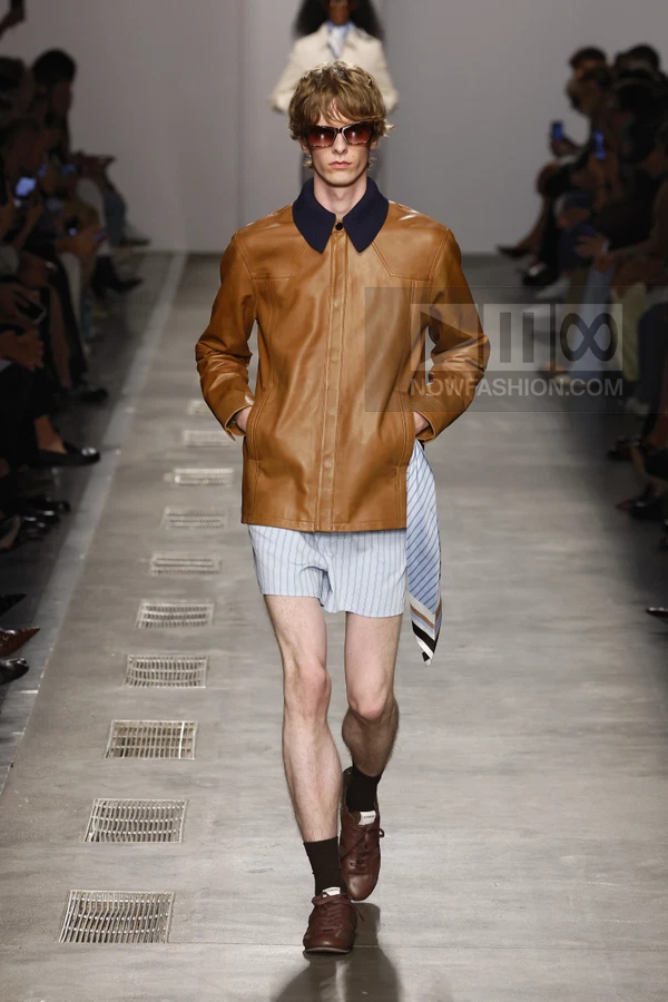 Model walking on runway wearing a tan leather jacket, light blue striped shorts, and brown sneakers with dark socks.