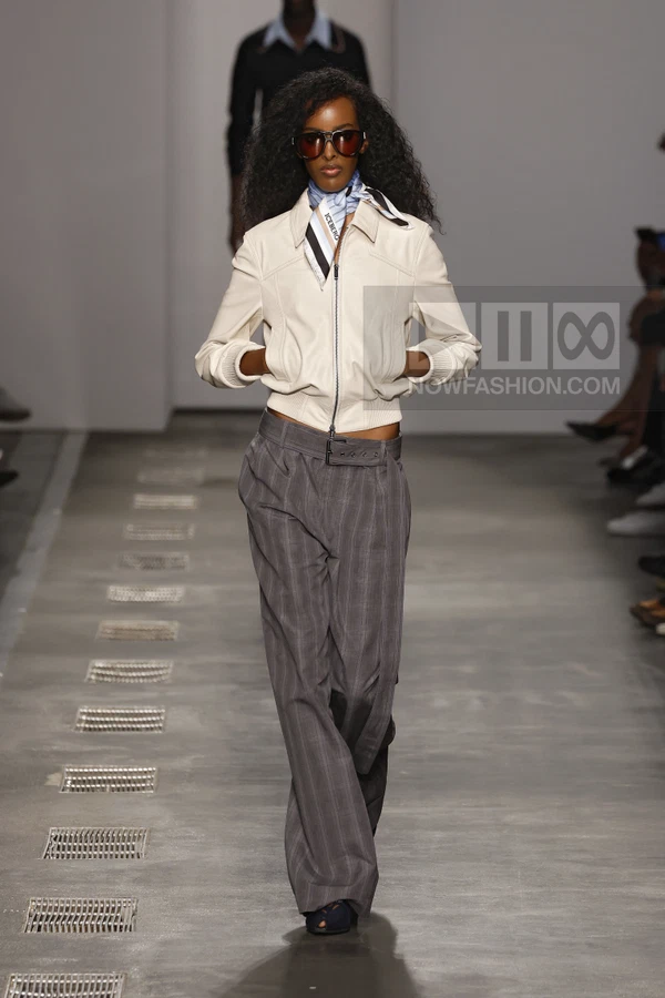Model wearing a white leather bomber jacket, gray pinstripe trousers, and a printed neck scarf on a runway.