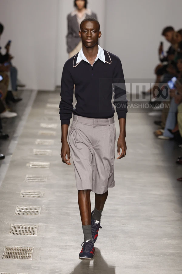 A person walks down a runway wearing a dark navy long-sleeve polo shirt with a light blue collar and gray tailored shorts.