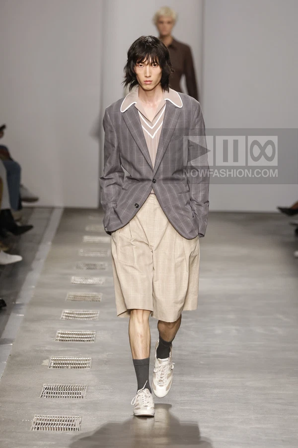 A person walks on a runway wearing a gray plaid blazer, beige pleated shorts, and white sneakers with gray socks.