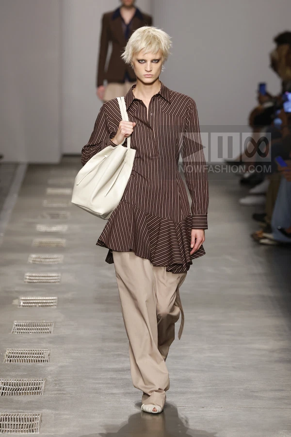 Model walking in a brown striped shirt with a ruffled hem, beige trousers, and a white shoulder bag on a runway.