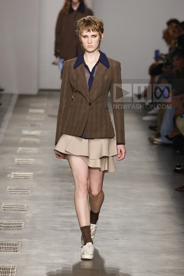 Model walking the runway in a brown pinstripe blazer, navy shirt, and beige tiered mini skirt with sneakers and socks.