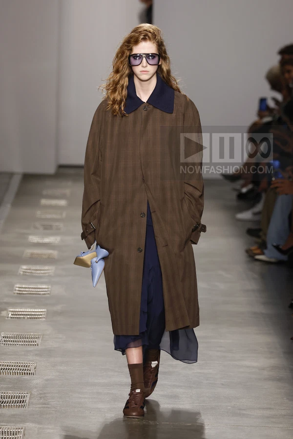 Model walking in a brown plaid oversized coat over a navy blue dress, brown sneakers, and holding light blue heels.