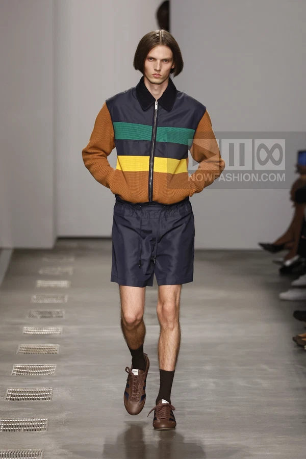 A person walks down a runway wearing a color-blocked zip-up jacket, navy shorts, brown sneakers, and dark socks.