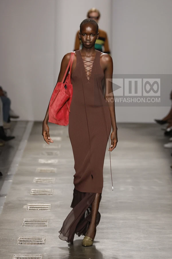 A model walks the runway in a brown ribbed midi dress with lace-up neckline, paired with a bright red tote bag.