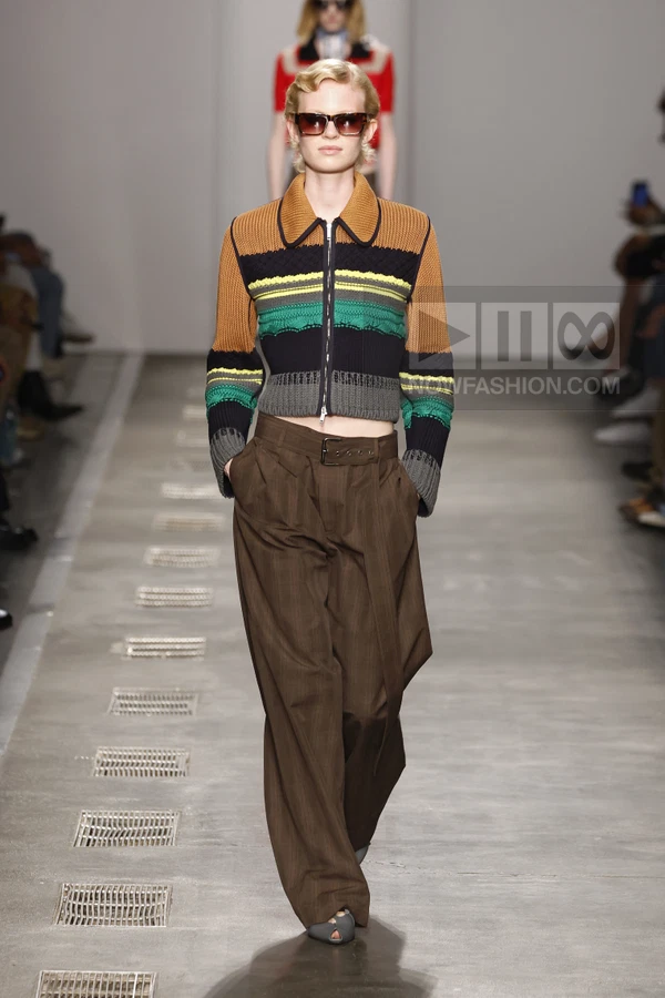 A model walks the runway wearing a multi-colored striped zip-up knit jacket and wide-leg brown trousers.