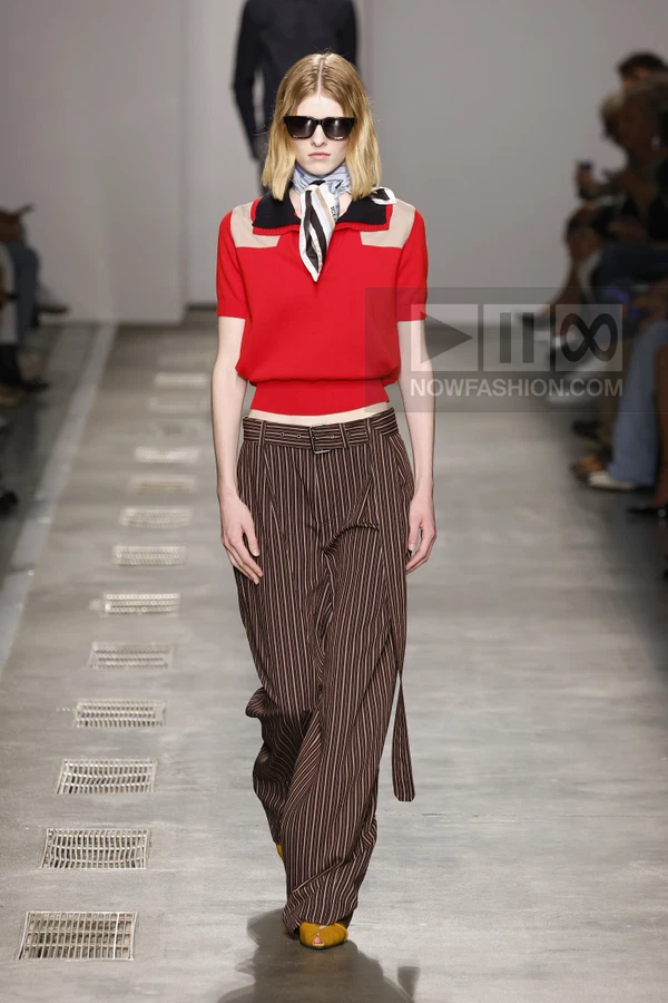 Model wearing a red polo top, brown pinstripe wide-leg trousers, a neck scarf, and sunglasses on a runway.