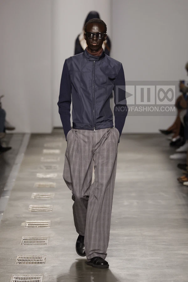 A person walks down a runway wearing a navy zip-up jacket with ribbed sleeves and gray plaid wide-leg trousers.