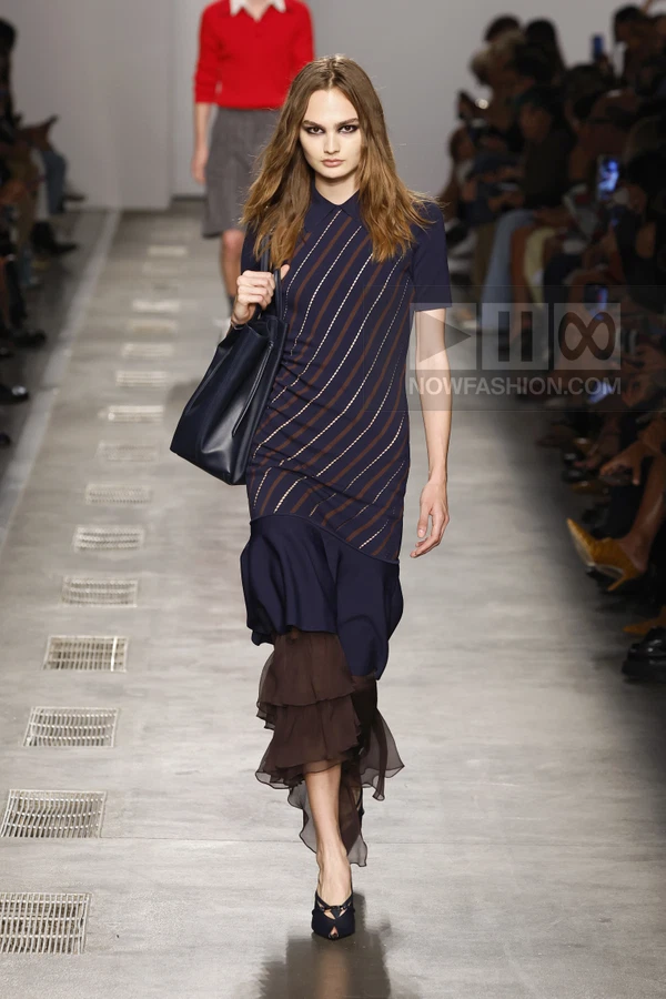 Model walking in a navy blue midi dress with diagonal stripes and a layered brown ruffled hem, carrying a navy tote bag.